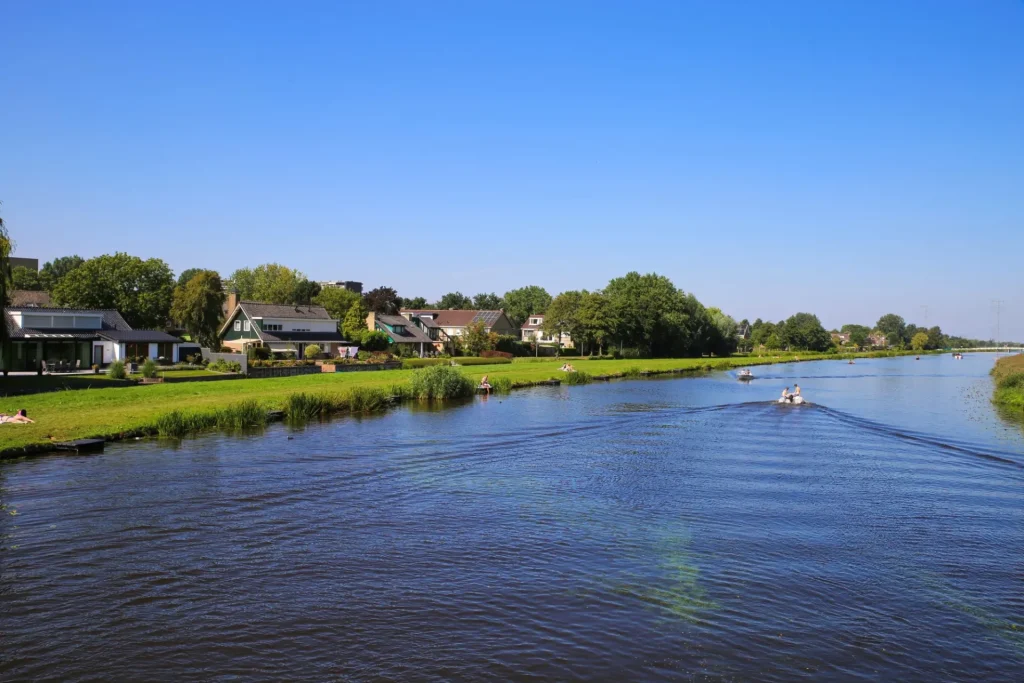 Blick über den Fluss Alblas auf eine Wiese mit Wohnhäusern vor blauem wolkenlosen Sommerhimmel - Alblasserdam, Niederlande