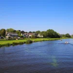 Blick über den Fluss Alblas auf eine Wiese mit Wohnhäusern vor blauem wolkenlosen Sommerhimmel - Alblasserdam, Niederlande